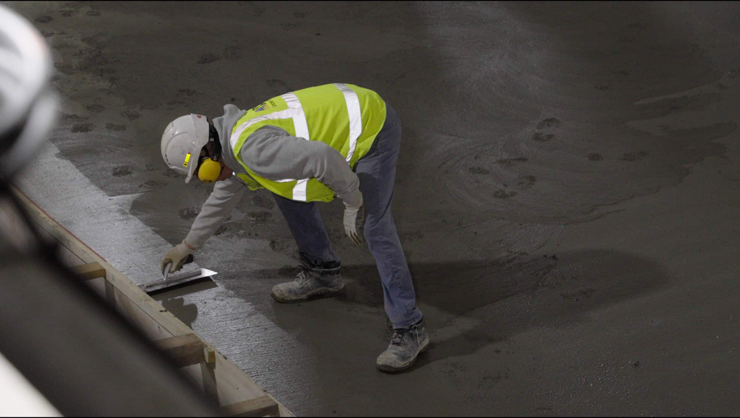 A concrete specialist with a high vis vest and white hard hat spreading concrete