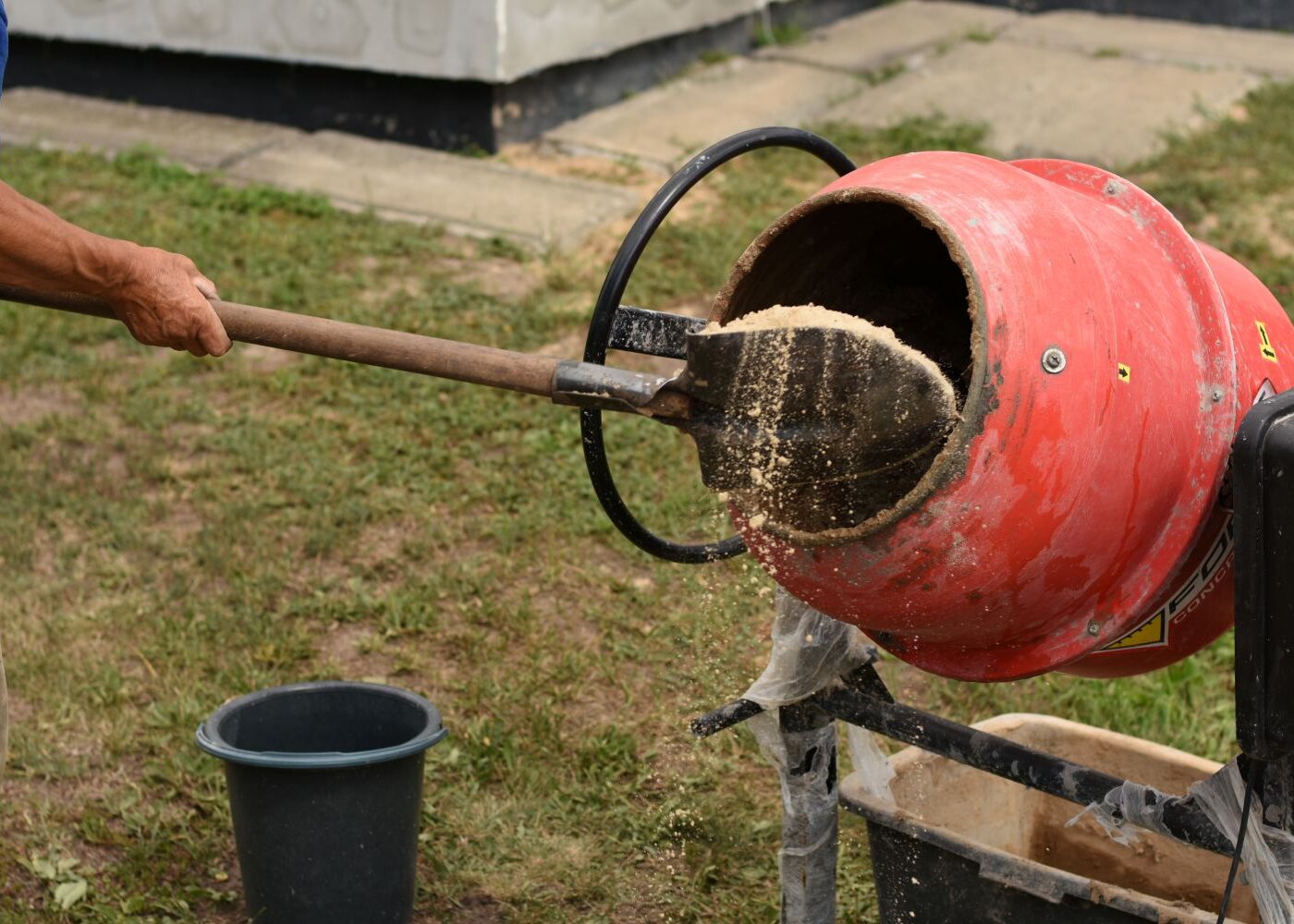 Concrete being mixed in a round, red mixer. There is a shovel adding sand into the mixture.