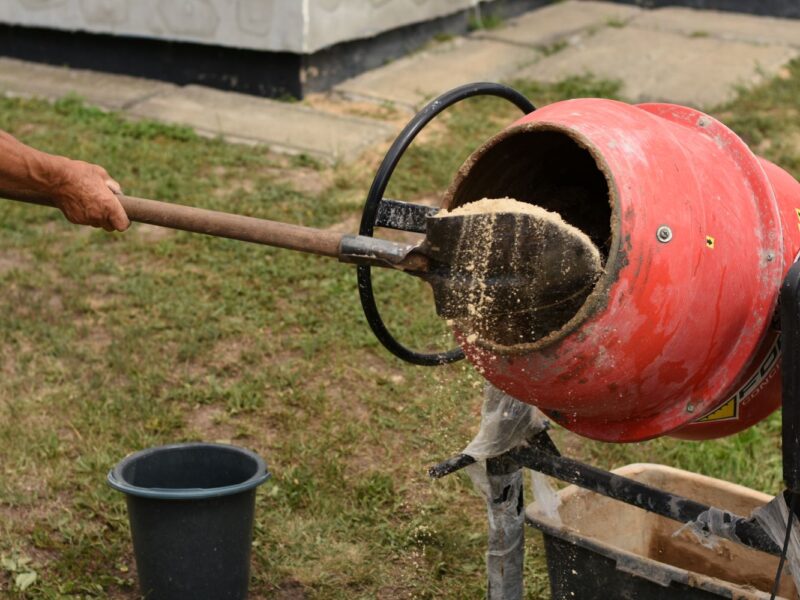 Concrete being mixed in a round, red mixer. There is a shovel adding sand into the mixture.